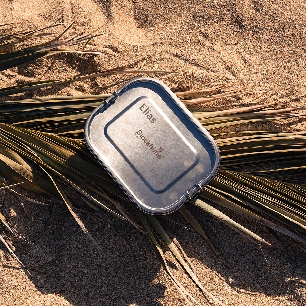 Stainless steel lunch box with the name Elias engraved into the lid, lying on sand between palm fronds