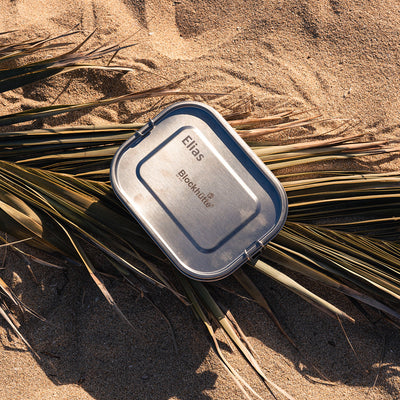 Stainless steel lunch box with the name Elias engraved into the lid, lying on sand between palm fronds