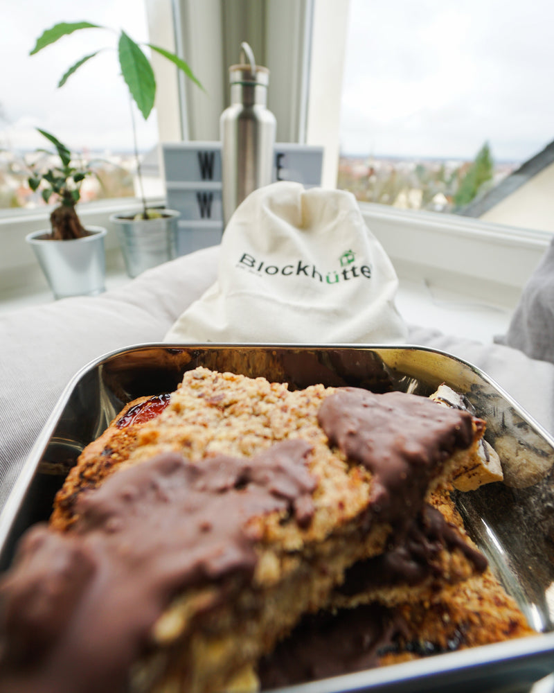 Open stainless steel lunch box filled with chocolate-topped cake bars, with a cotton bag and a steel bottle on the windowsill behind