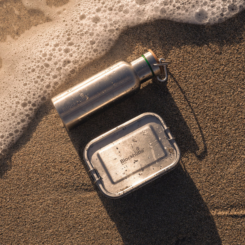 Stainless steel water bottle and lunch box lying in wet sand at the water's edge