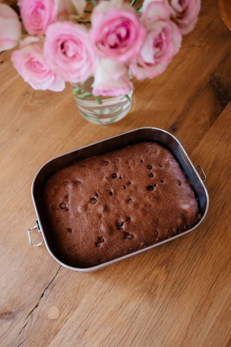 Chocolate cake baked in a stainless steel lunch box, cooling on a wooden table next to a vase of pink roses