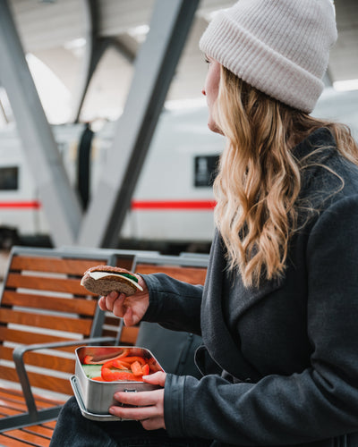 A woman on a station bench eating a sandwich from an open stainless steel lunch box with sliced peppers and cucumber