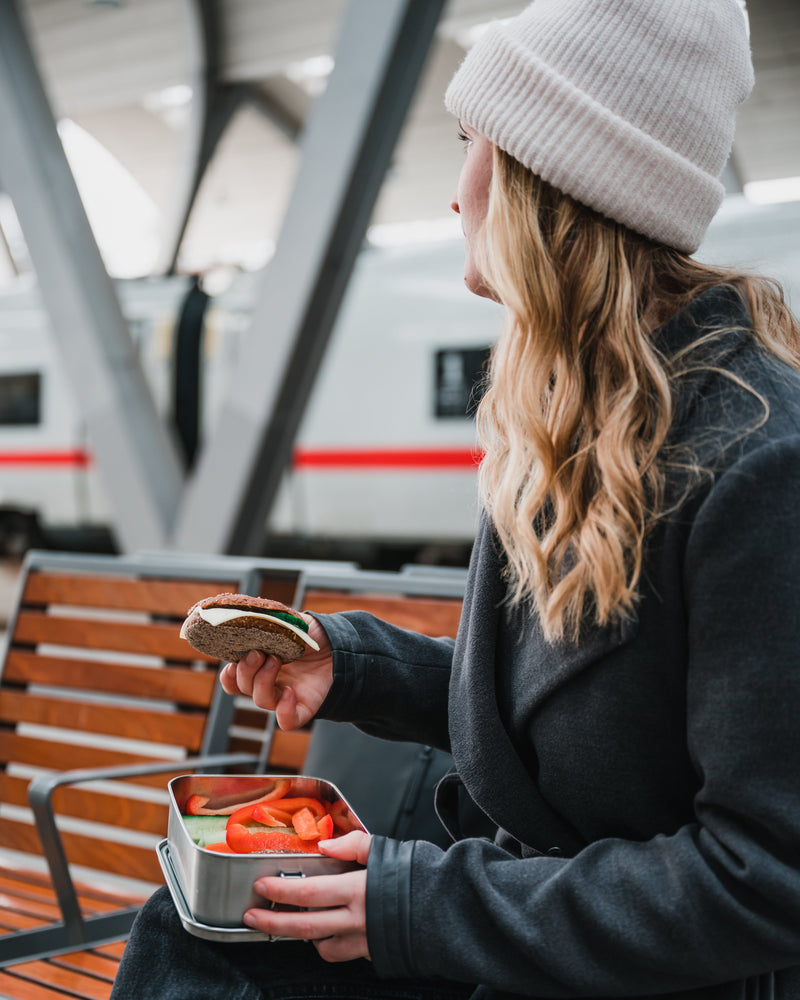 A woman on a station bench eating a sandwich from an open stainless steel lunch box with sliced peppers and cucumber
