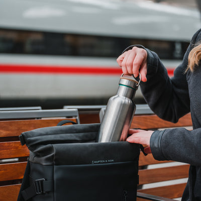 Stainless steel water bottle with a bamboo cap being slid into a black backpack on a station bench