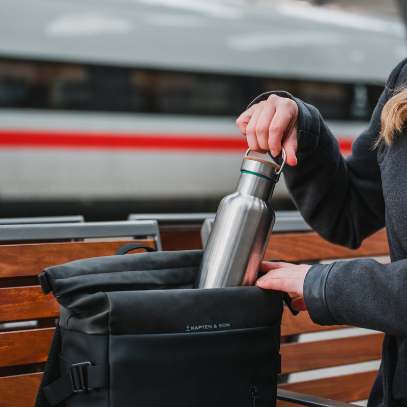 Stainless steel water bottle with a bamboo cap being slid into a black backpack on a station bench
