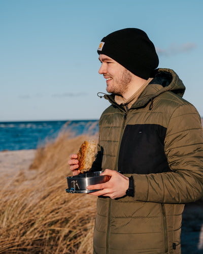 A man eating a sandwich out of a Blockhuette stainless steel lunch box at the coast