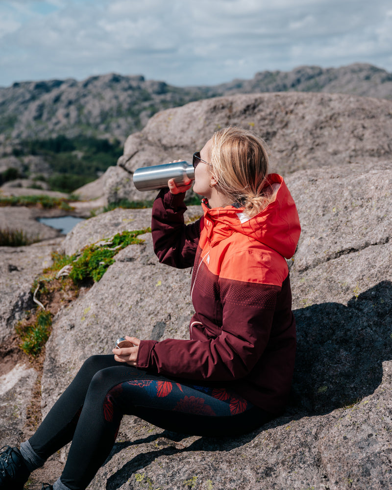 A woman in a red jacket sitting on a rock, drinking from a stainless steel water bottle