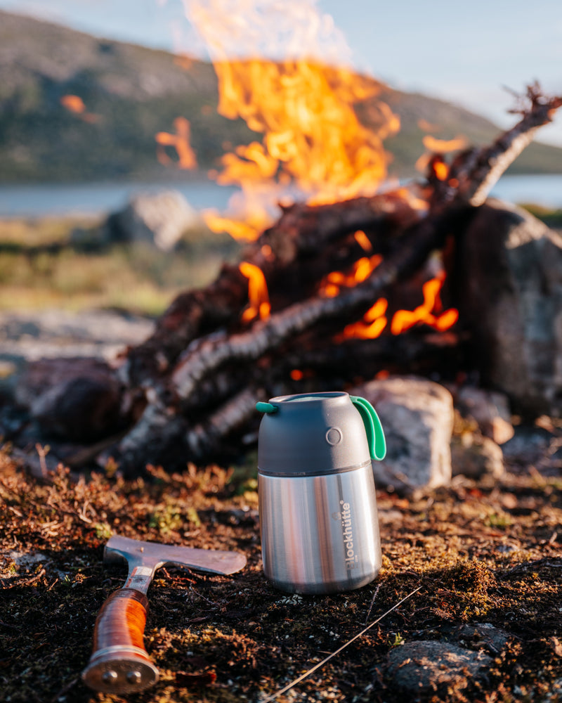 Insulated food jar standing in front of a campfire next to an axe, with a lake and hills behind