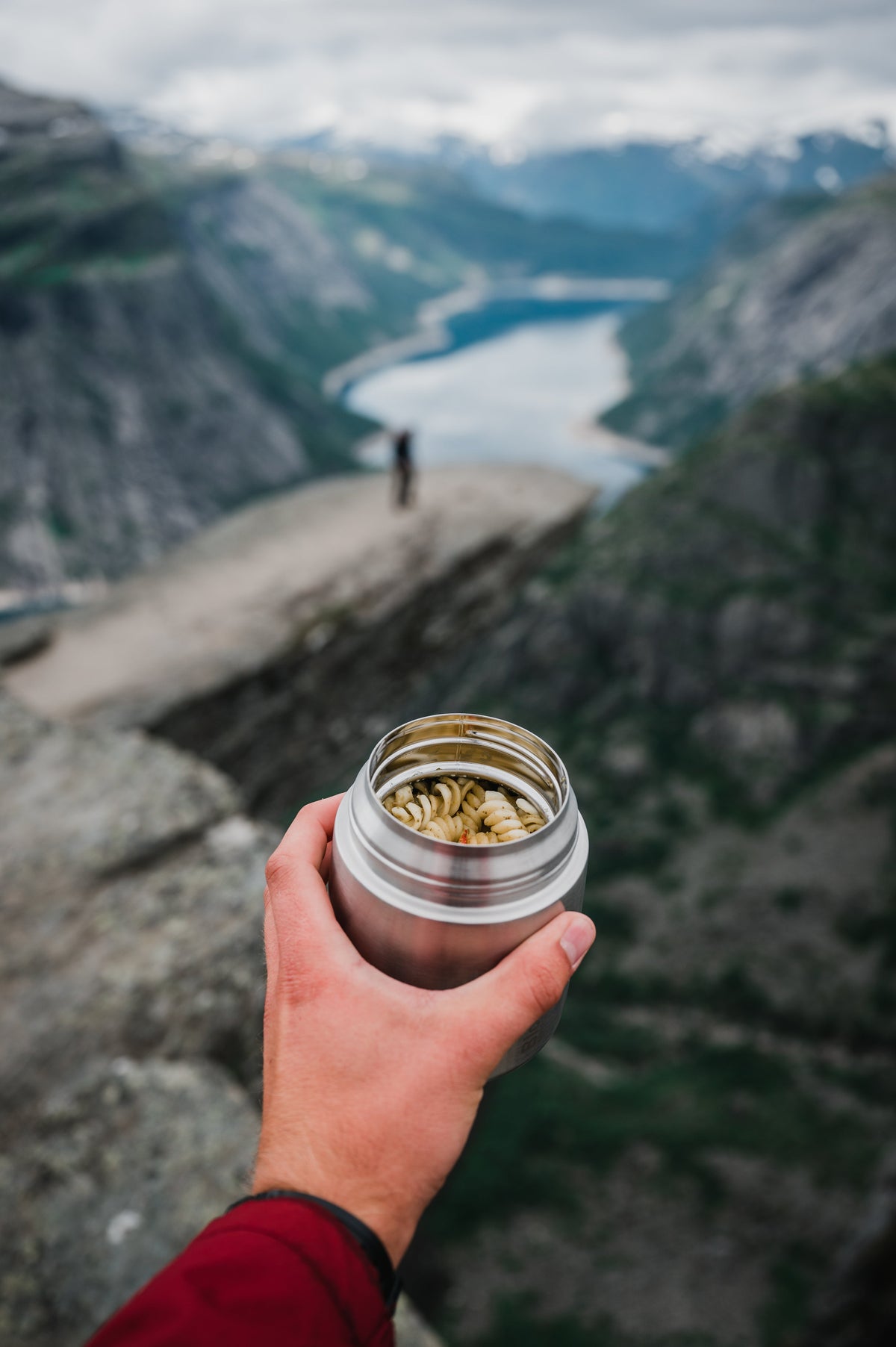 Open insulated food jar with pasta, held in one hand above a fjord