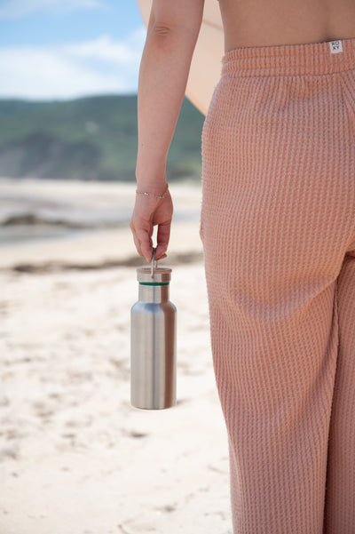 Stainless steel water bottle with a bamboo cap, held by its loop at the beach