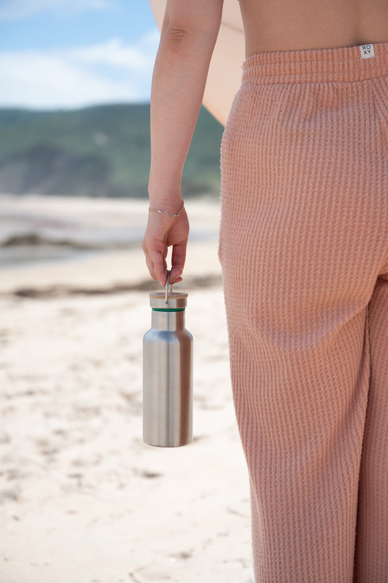 Stainless steel water bottle with a bamboo cap, held by its loop at the beach