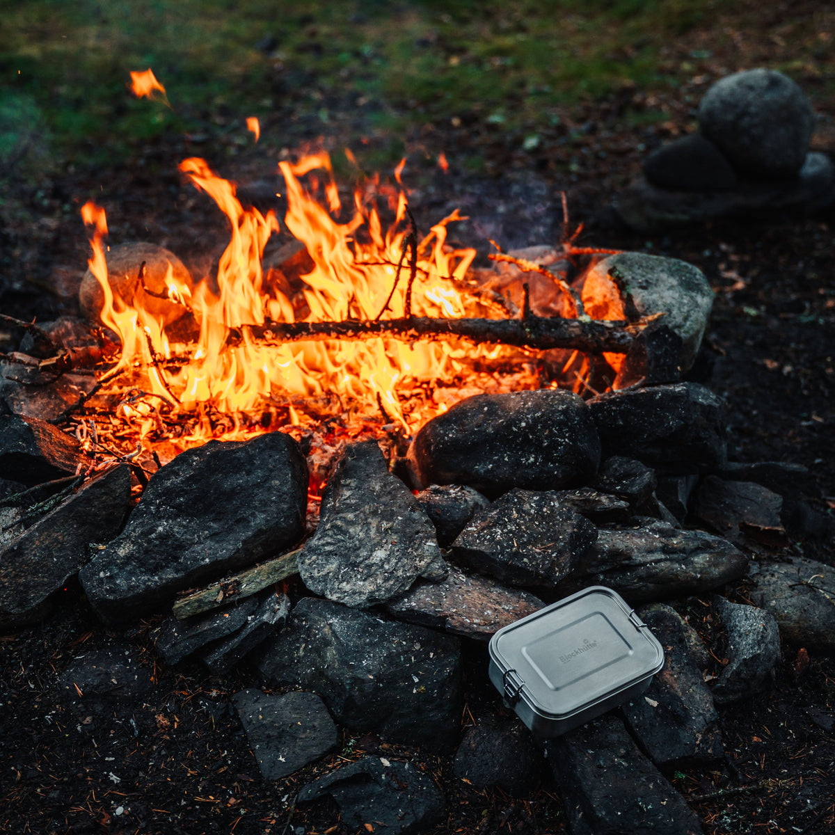 Stainless steel lunch box standing on stones next to a campfire