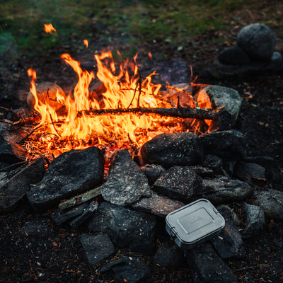 Stainless steel lunch box standing on stones next to a campfire