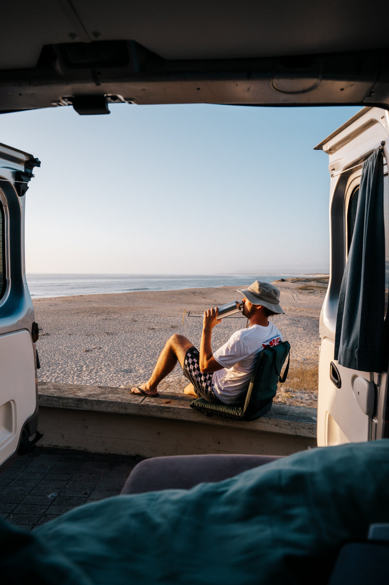 A man drinking from a Blockhuette stainless steel water bottle outside a van at the beach