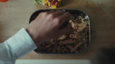 Still from the film Beyond the Lunchbox: a hand filling the stainless steel lunch box with rice and chicken