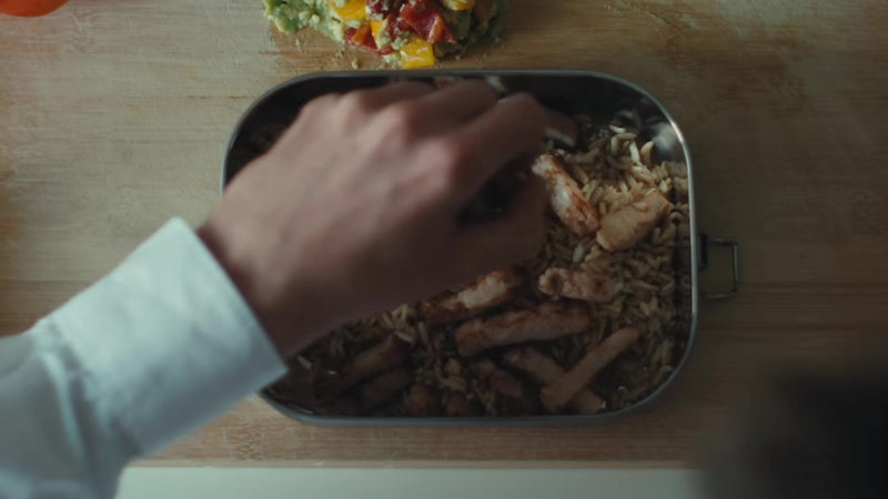 Still from the film Beyond the Lunchbox: a hand filling the stainless steel lunch box with rice and chicken