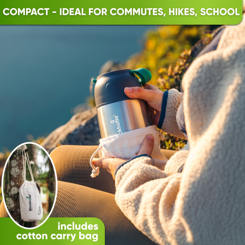 Person in a fleece jacket pulling a steel food jar with dark lid and green strap from its cotton bag beside a lake
