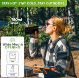 Woman in sunglasses and a checked fleece jacket drinking from the 25 oz bottle in a black silicone sleeve at a forest picnic table