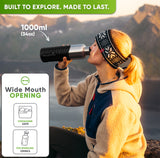 Woman in a knit headband drinking from the 34 oz bottle in a black silicone sleeve on a mountain at sunset