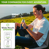 Smiling man sitting on a grassy hillside above the sea, holding the 17 oz bottle in a blue silicone sleeve