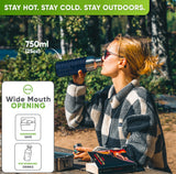 Woman in sunglasses and a checked fleece jacket drinking from the 25 oz bottle in a blue silicone sleeve at a forest picnic table
