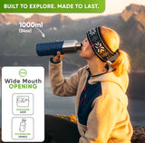 Woman in a knit headband drinking from the 34 oz bottle in a blue silicone sleeve on a mountain at sunset