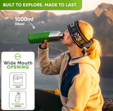 Woman in a knit headband drinking from the 34 oz bottle in a bright green silicone sleeve on a mountain at sunset