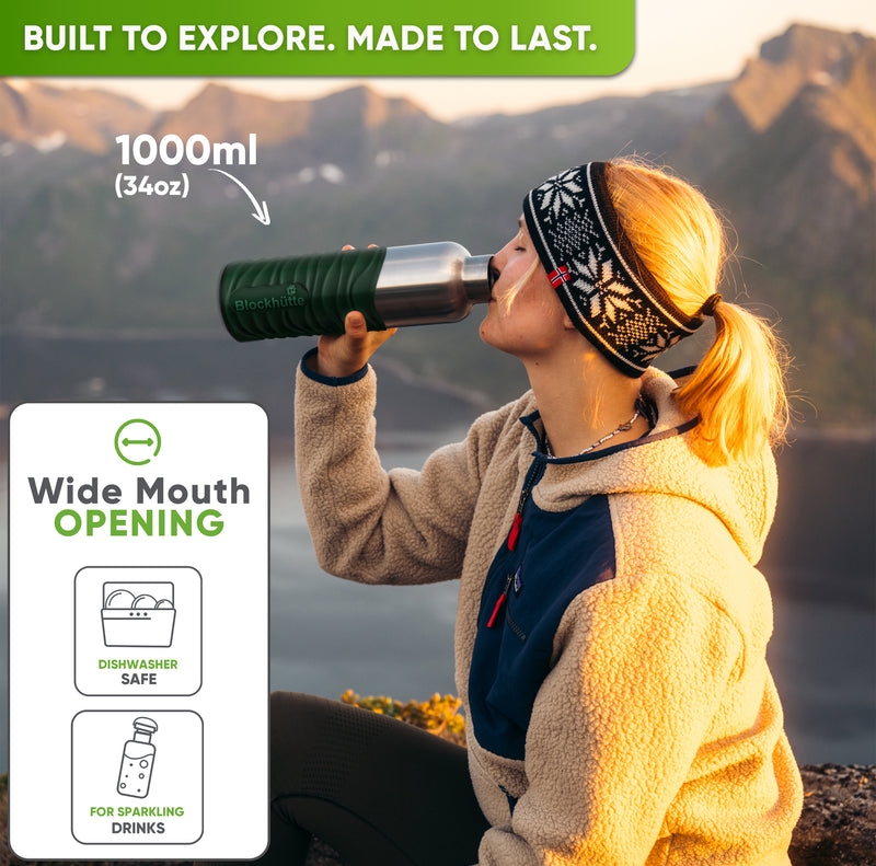Woman in a knit headband drinking from the 34 oz bottle in a dark green silicone sleeve on a mountain at sunset