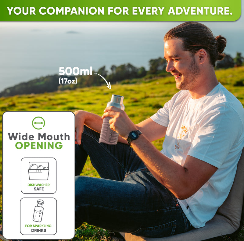 Smiling man sitting on a grassy hillside above the sea, holding the 17 oz bottle in a pink silicone sleeve