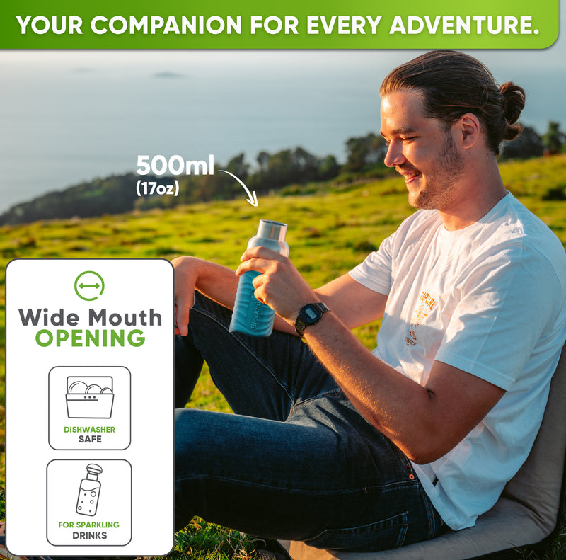 Smiling man sitting on a grassy hillside above the sea, holding the 17 oz bottle in a turquoise silicone sleeve