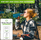 Woman in sunglasses and a checked fleece jacket drinking from the 25 oz bottle in a turquoise silicone sleeve at a forest picnic table