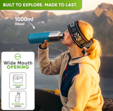 Woman in a knit headband drinking from the 34 oz bottle in a turquoise silicone sleeve on a mountain at sunset