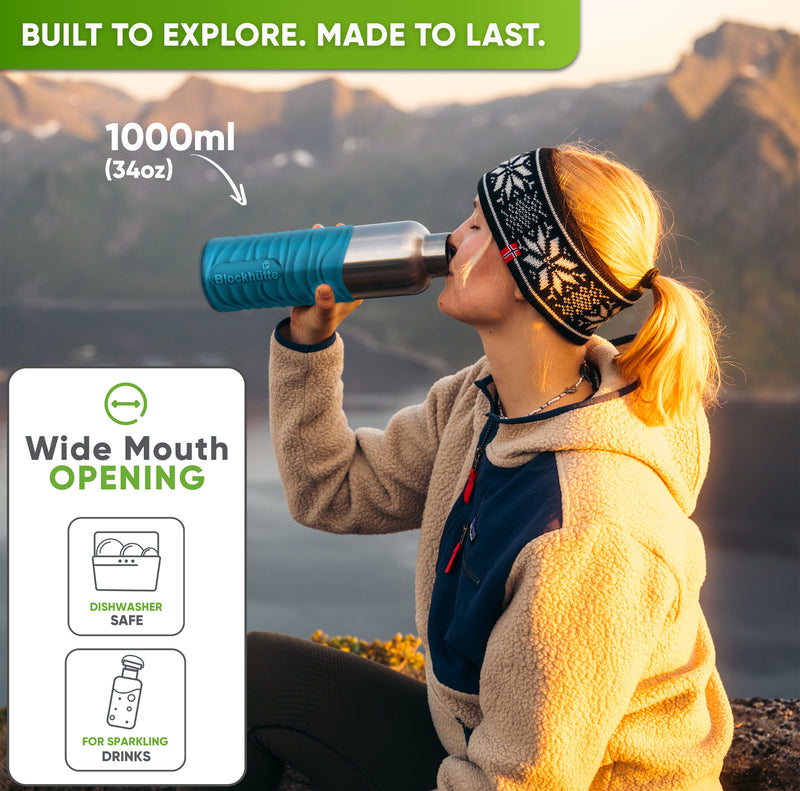 Woman in a knit headband drinking from the 34 oz bottle in a turquoise silicone sleeve on a mountain at sunset