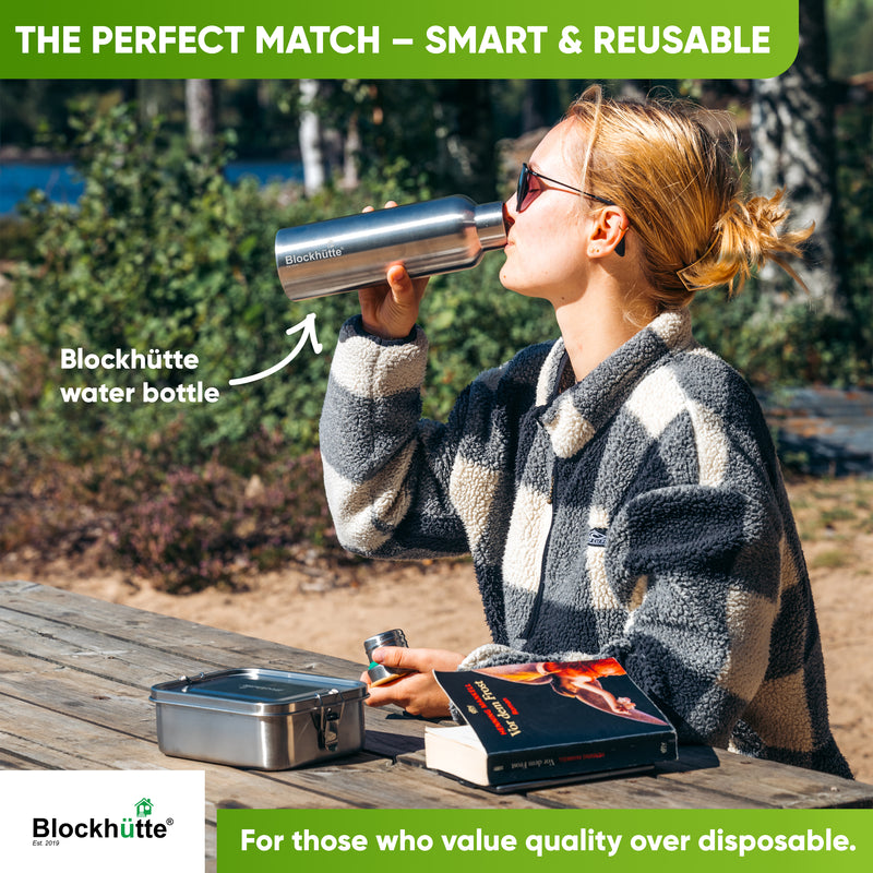 Woman in a fleece jacket drinking from a Blockhütte water bottle at a picnic table, closed lunch box and a book beside her
